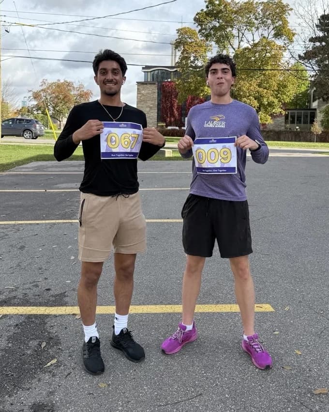 Jasman Mander and a friend holding race bibs 067 and 009 in a parking lot before a run.